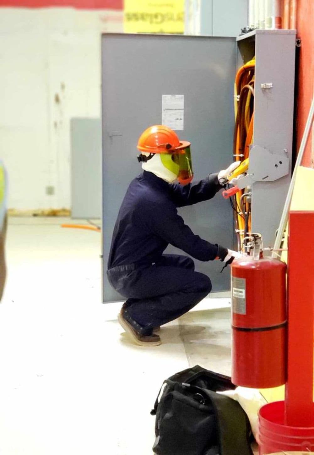 Electrician in safety gear operating electrical panel in industrial setting, fire extinguisher nearby.