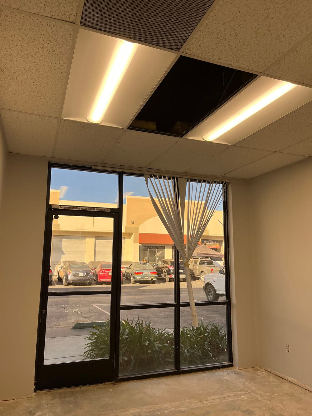 Office interior with damaged ceiling tiles, large windows, and view of parked cars outside.