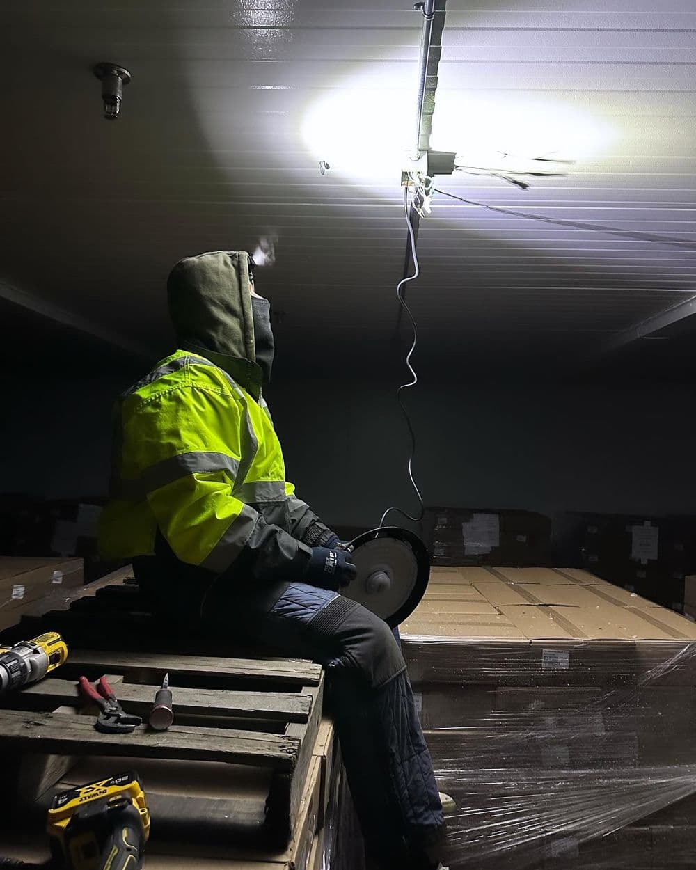 Worker in safety gear seated on pallets, repairing overhead lighting in a warehouse.