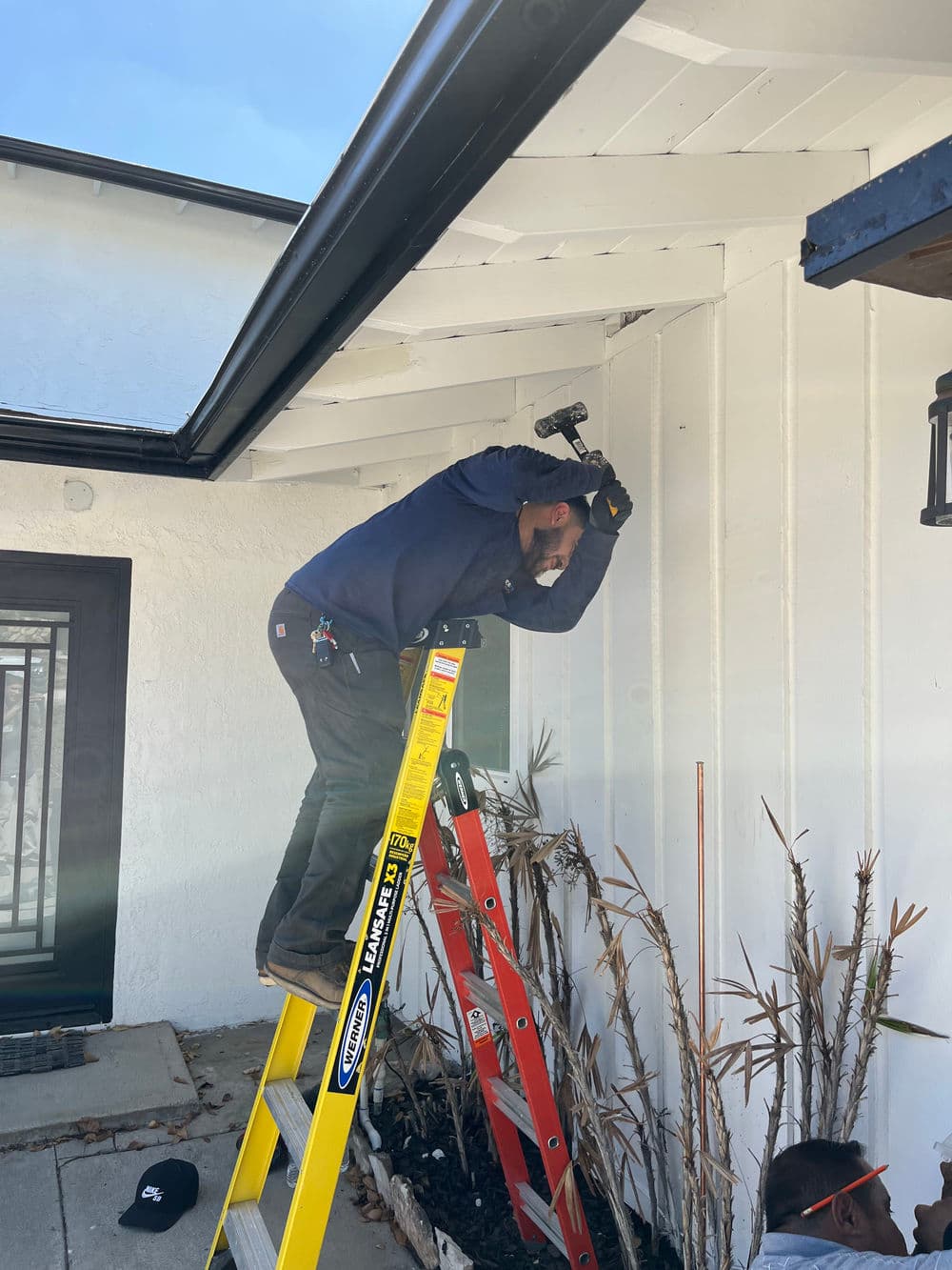 Worker repairing exterior with hammer while on ladder at residential property.