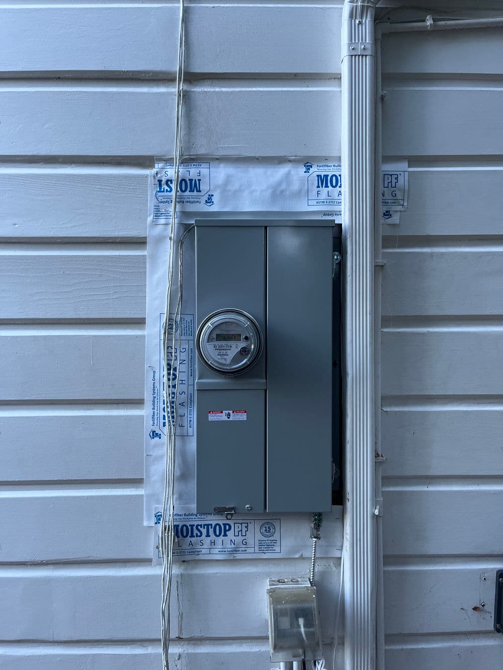 Electrical meter box mounted on a white wooden wall with moisture barrier flashing.