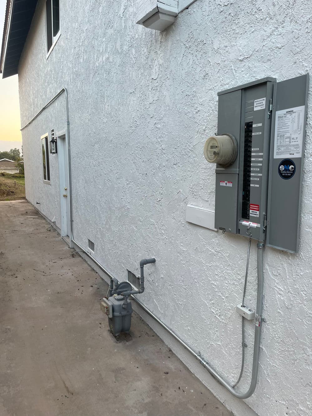 Utility panel and gas meter on the exterior wall of a white house in a residential area.