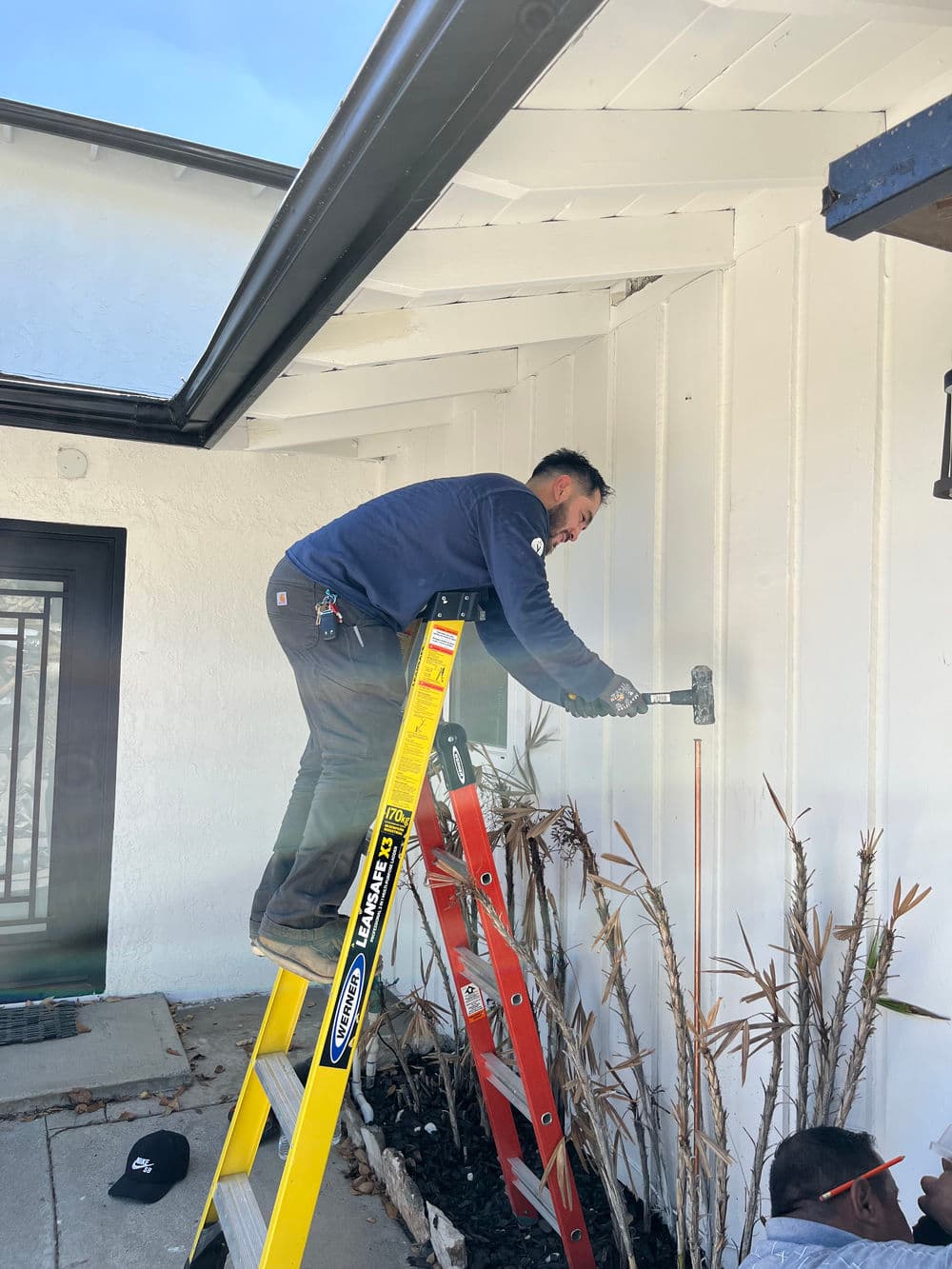 Worker on ladder installing fixtures on exterior wall of a house with manicured landscaping.