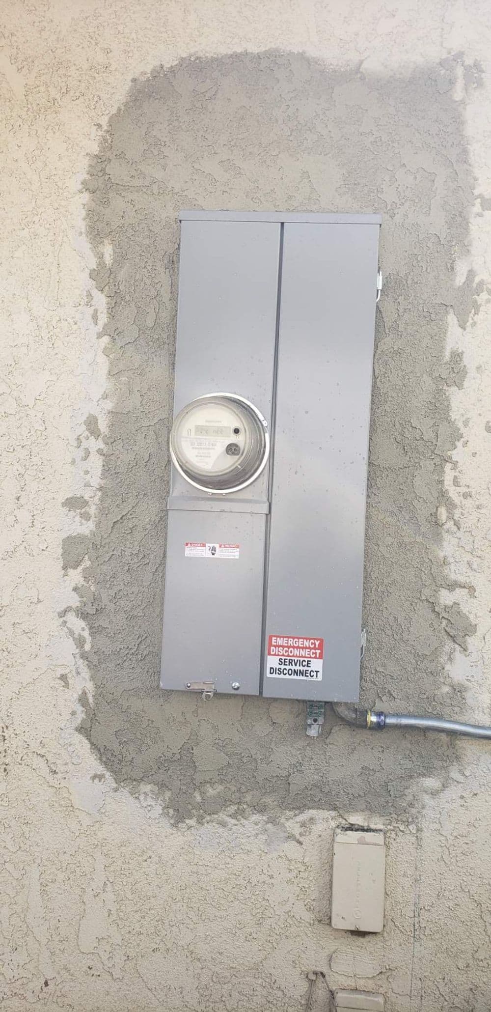 Gray electrical meter box on a textured wall with emergency disconnect label visible.