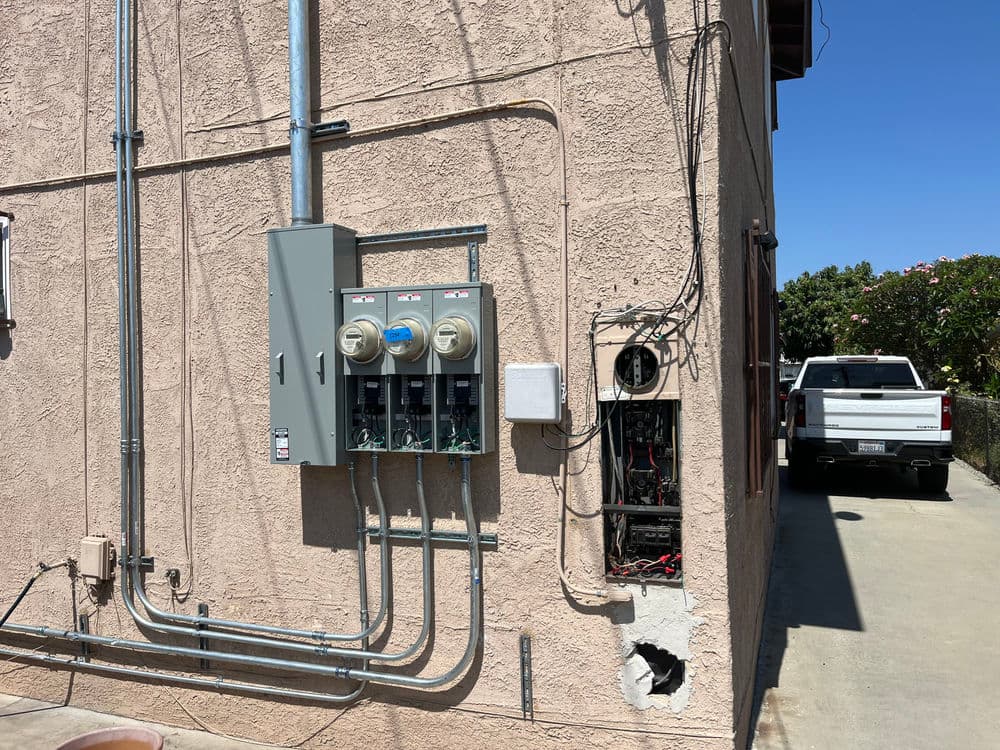 Electrical meters and panels on a light-colored wall beside a parking area.