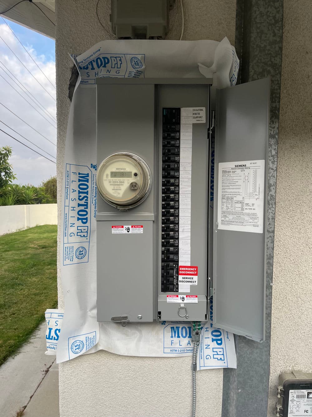 Electrical panel with meter, located outside a building, featuring safety labels and warnings.