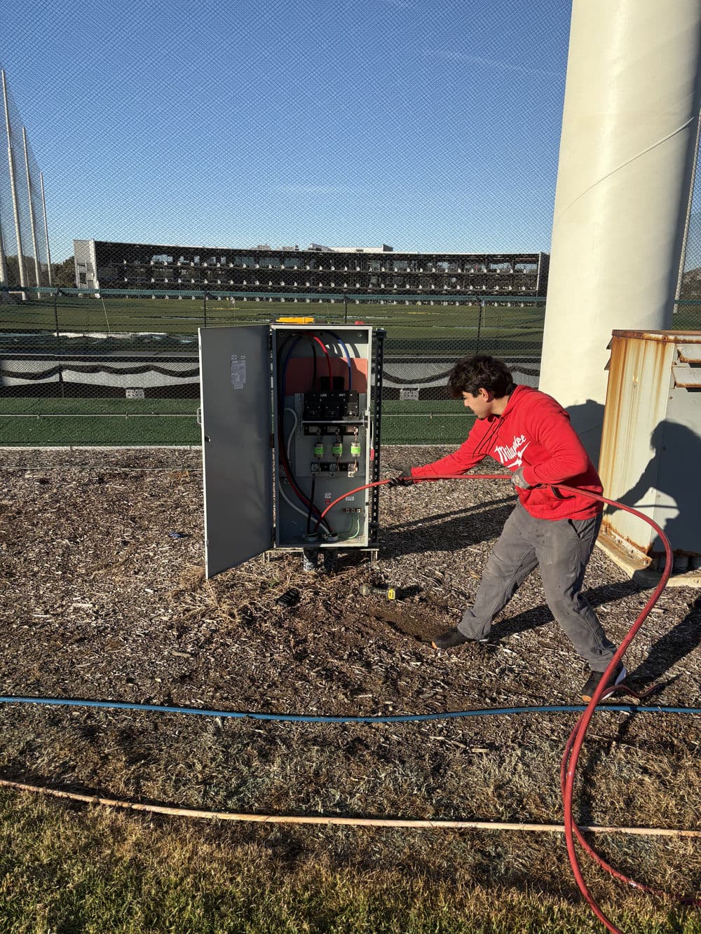 Technician servicing electrical equipment near a sports field with tools and hoses.