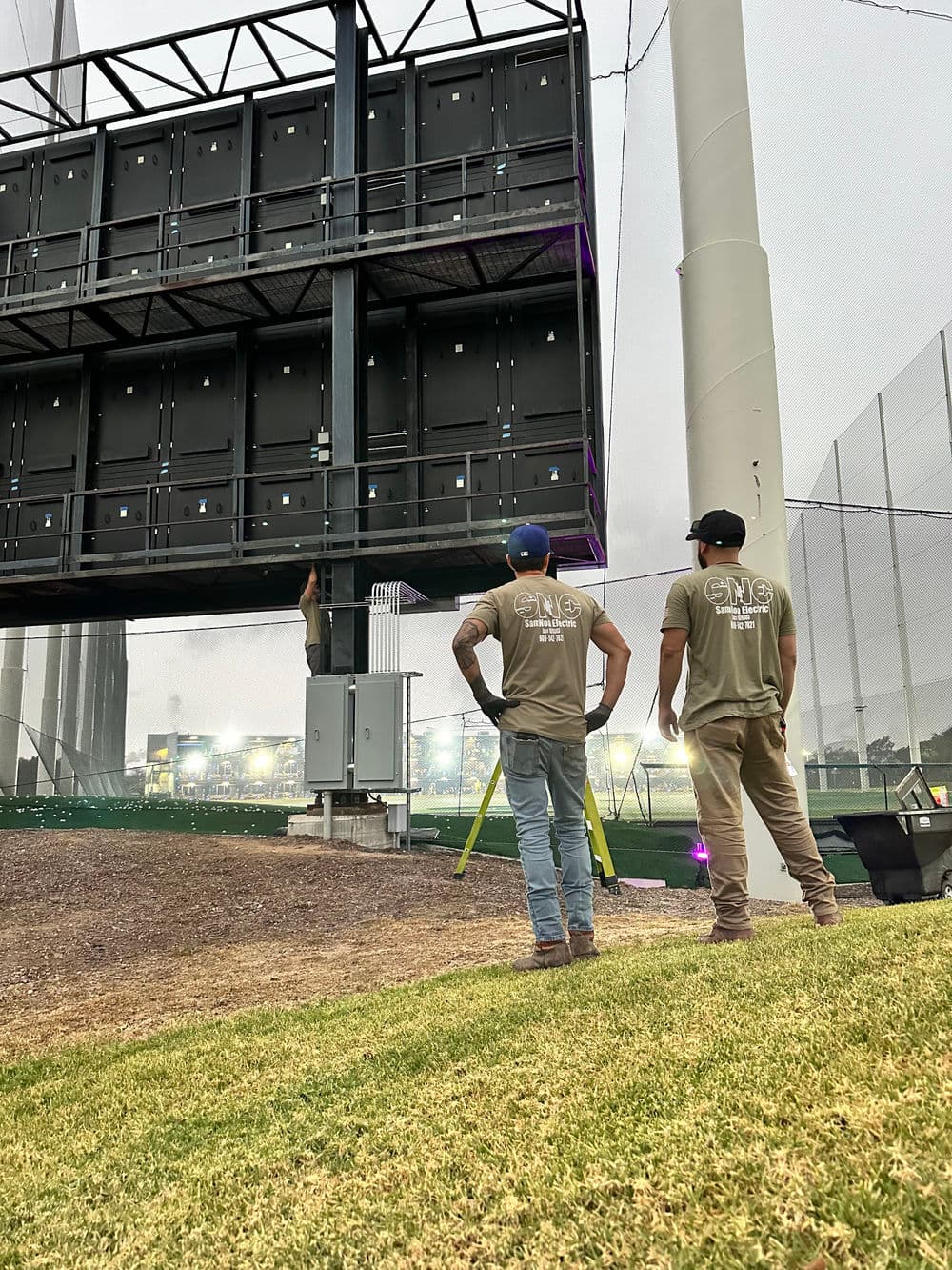 Workers adjusting a large electronic scoreboard at a sports facility during a cloudy day.
