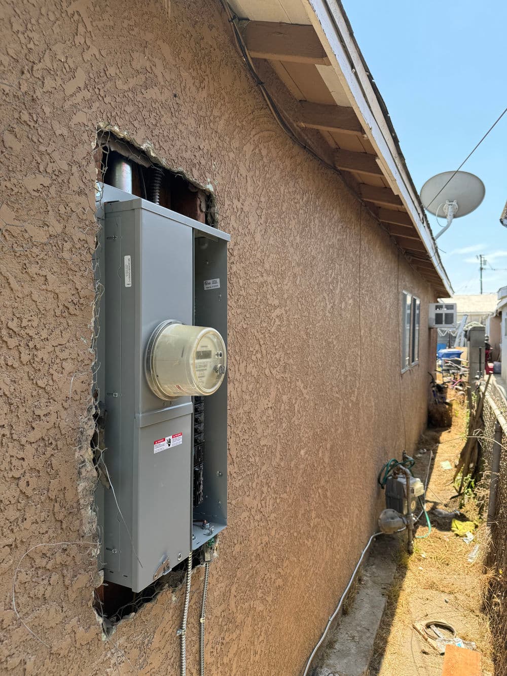 Electric meter installation on exterior wall of a house with satellite dish in background.