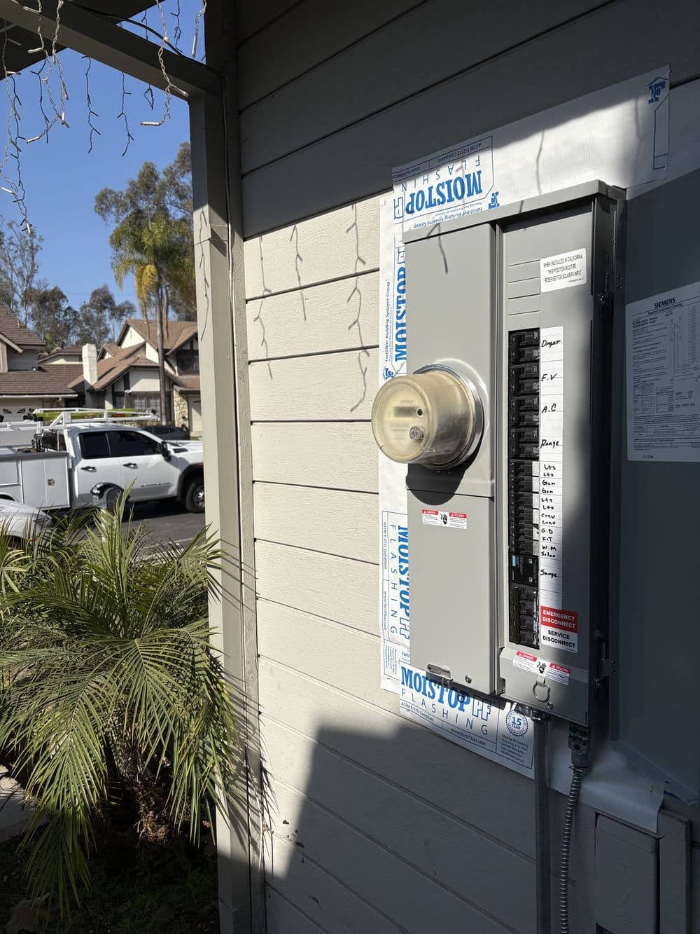 Electric meter mounted on a wall beside a circuit breaker box with white truck in the background.