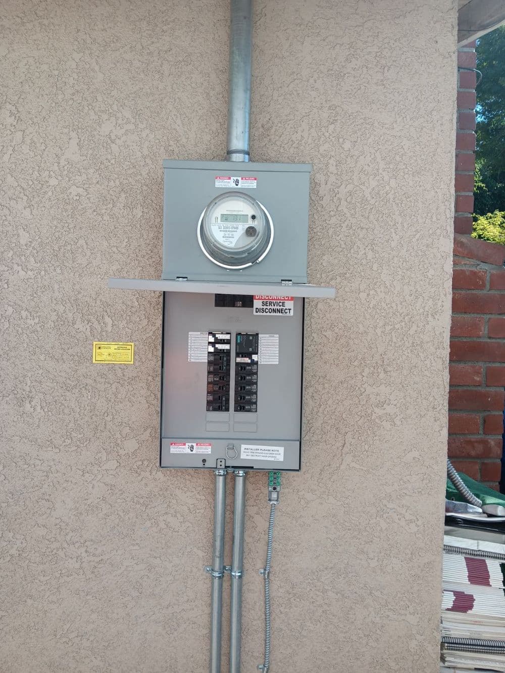 Electric meter and service panel mounted on a beige wall with safety labels.