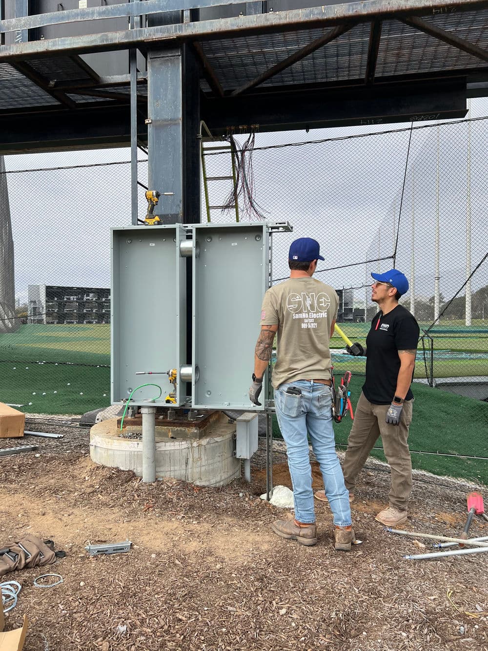 Technicians installing electrical equipment on a sports field structure.