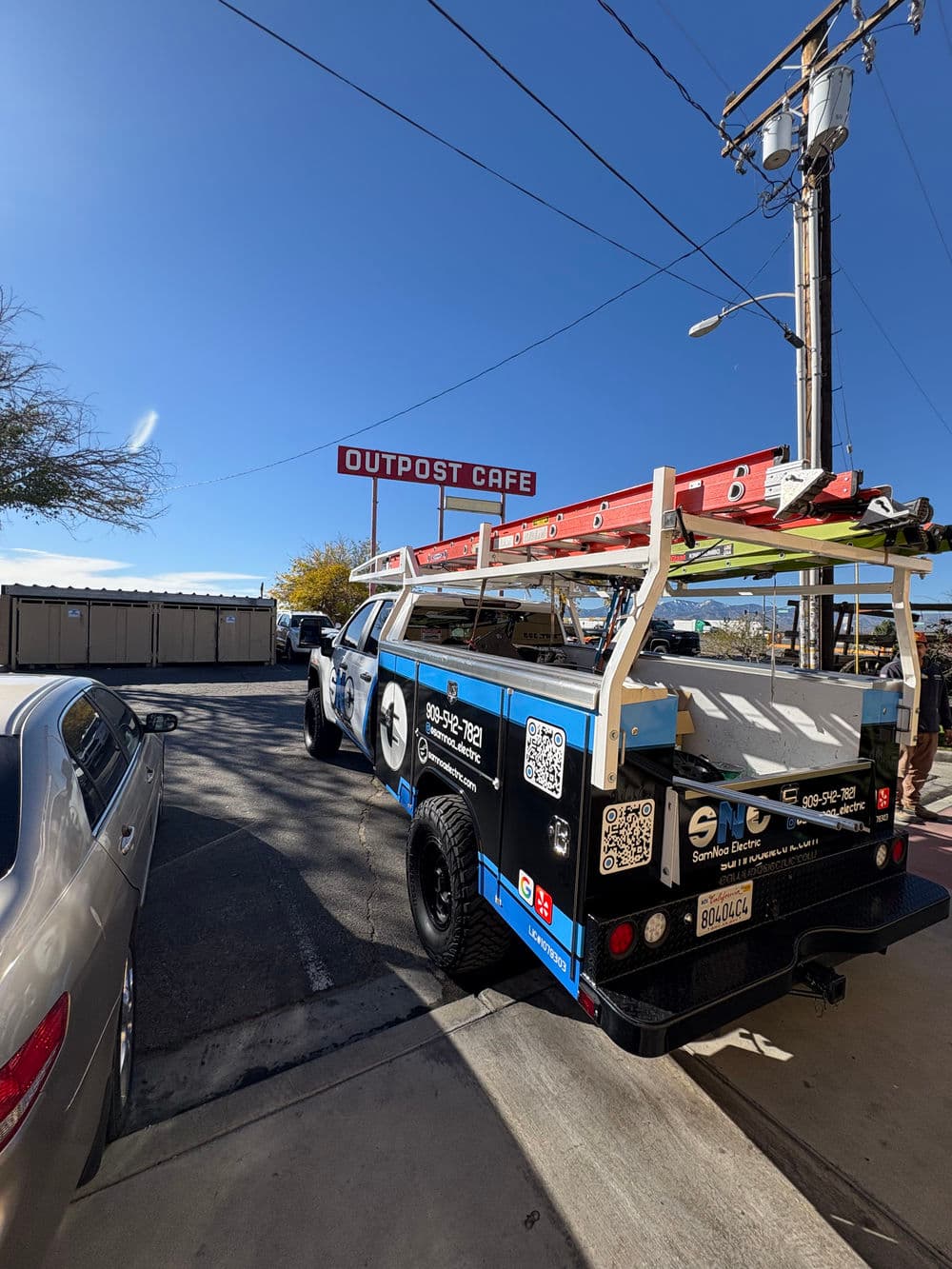 Service truck with ladders parked near Outpost Café under a clear blue sky.