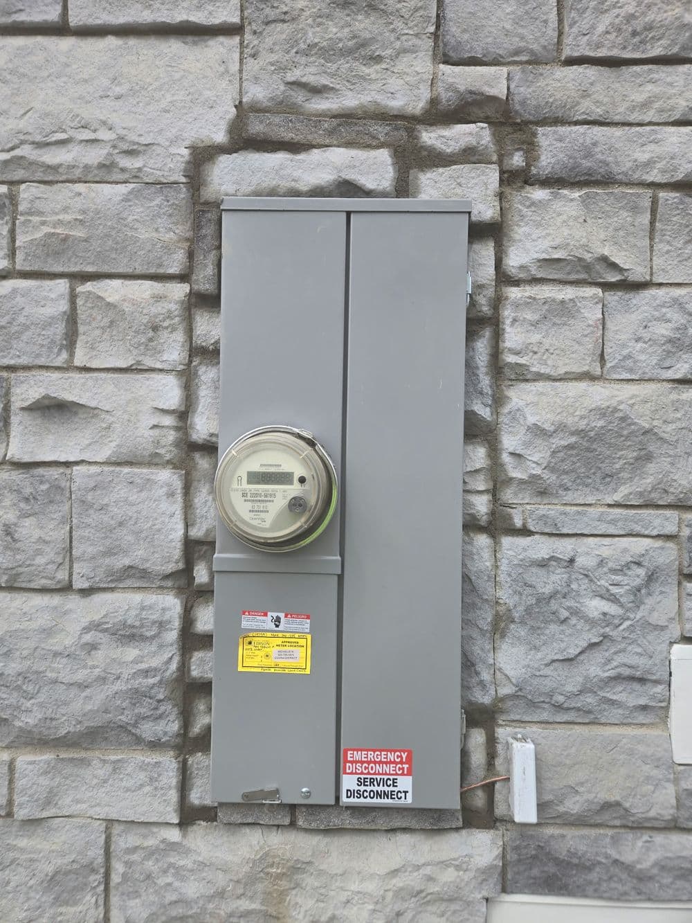 Electric meter mounted on a stone wall with emergency disconnect instructions.
