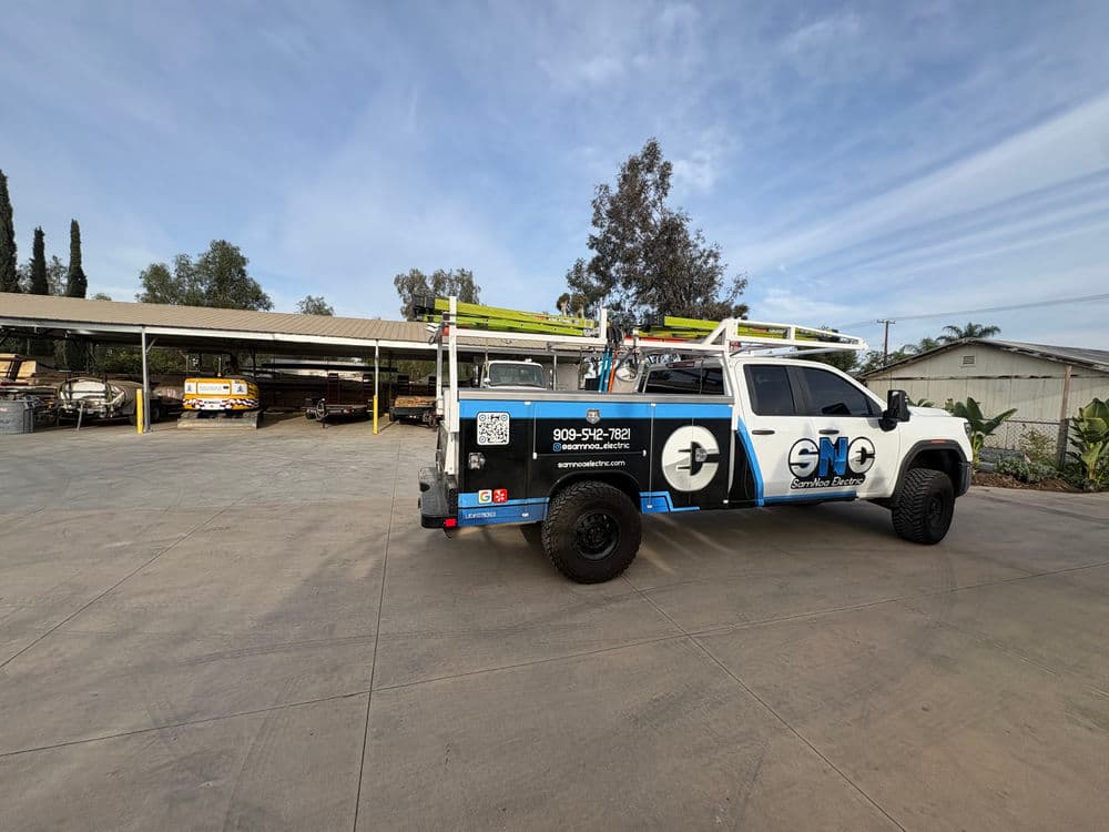 Service truck with equipment parked in a yard with storage buildings and trees in the background.