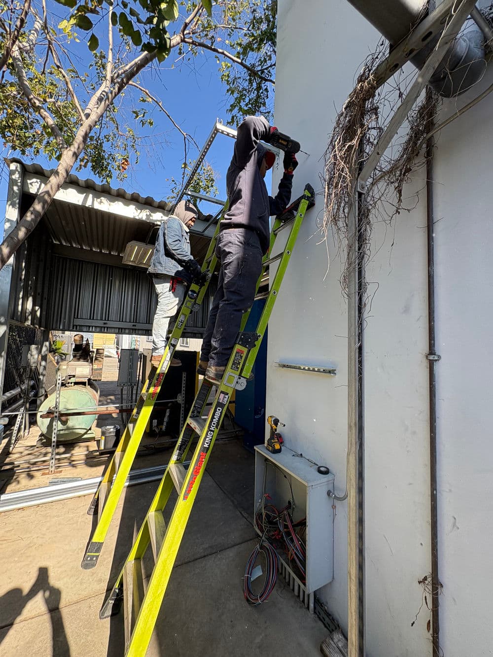 Worker on a ladder using a drill to install equipment on a building exterior.