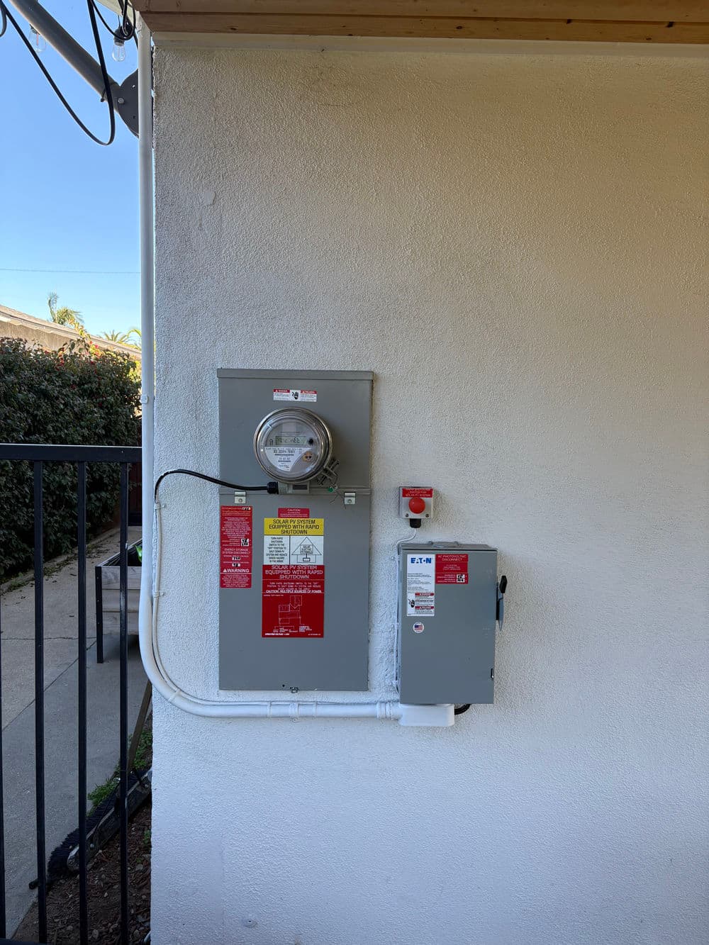 Electric meter and panel mounted on a white wall, surrounded by greenery and a walkway.