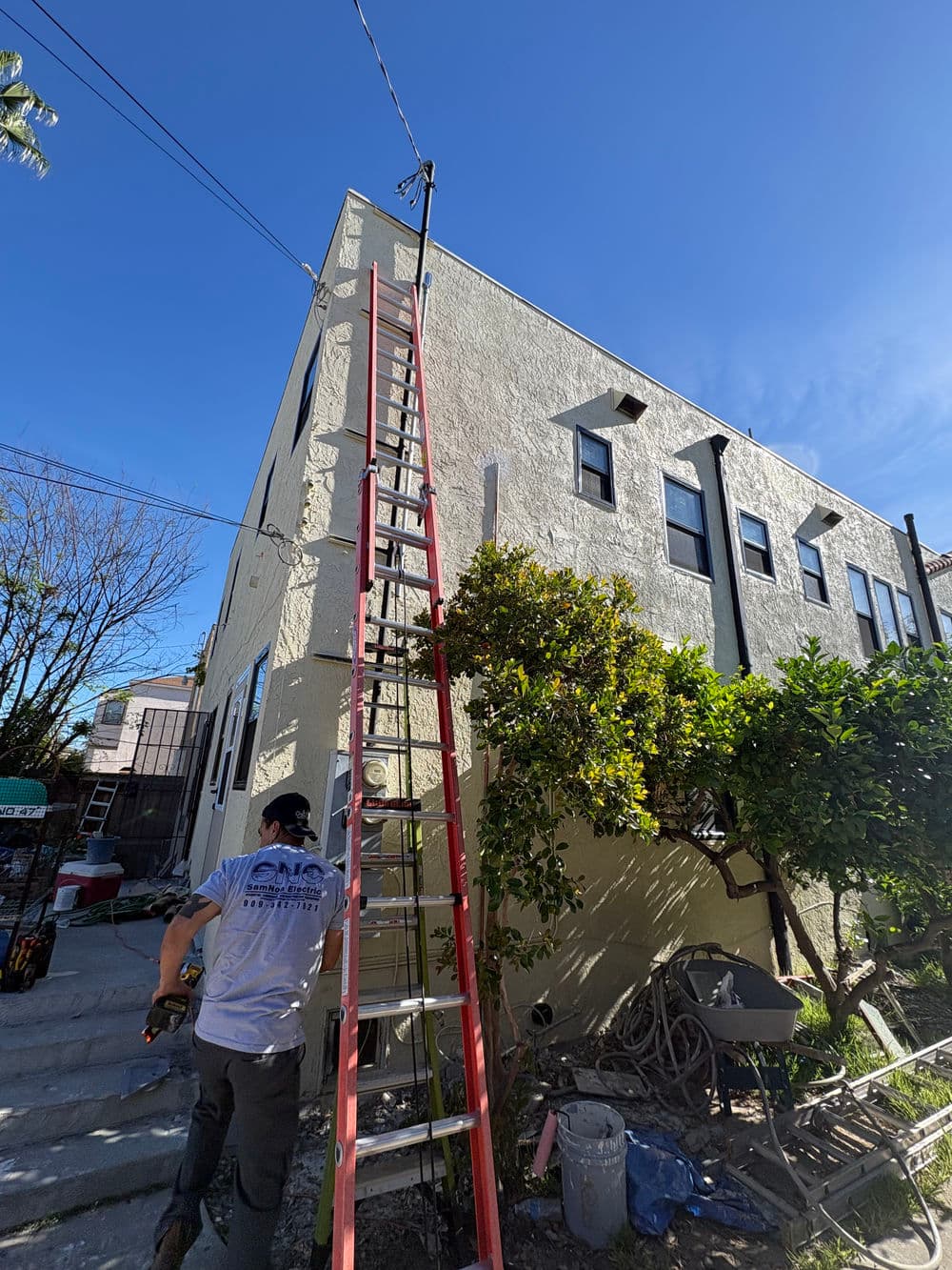 Worker using a tall ladder to perform maintenance on a two-story building under a clear blue sky.