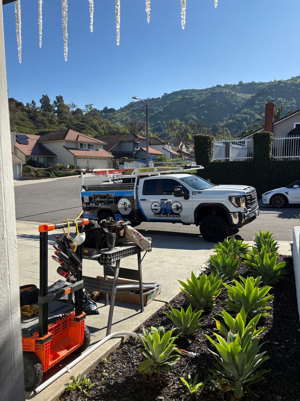 Service truck parked on a sunny street with hillside views and outdoor equipment nearby.